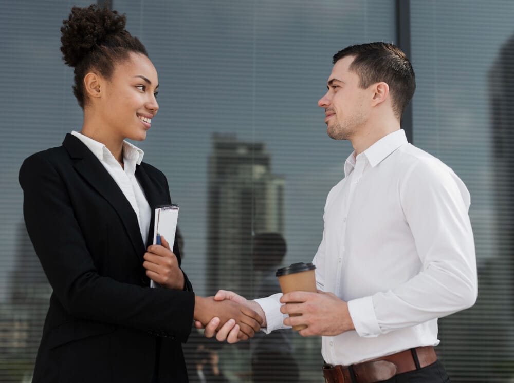 A businesswoman and a businessman shaking hands outdoors, with the businesswoman holding a notebook and the businessman holding a coffee cup. Both are smiling in a corporate environment.